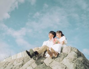 青空の下で岩に腰掛ける新郎新婦