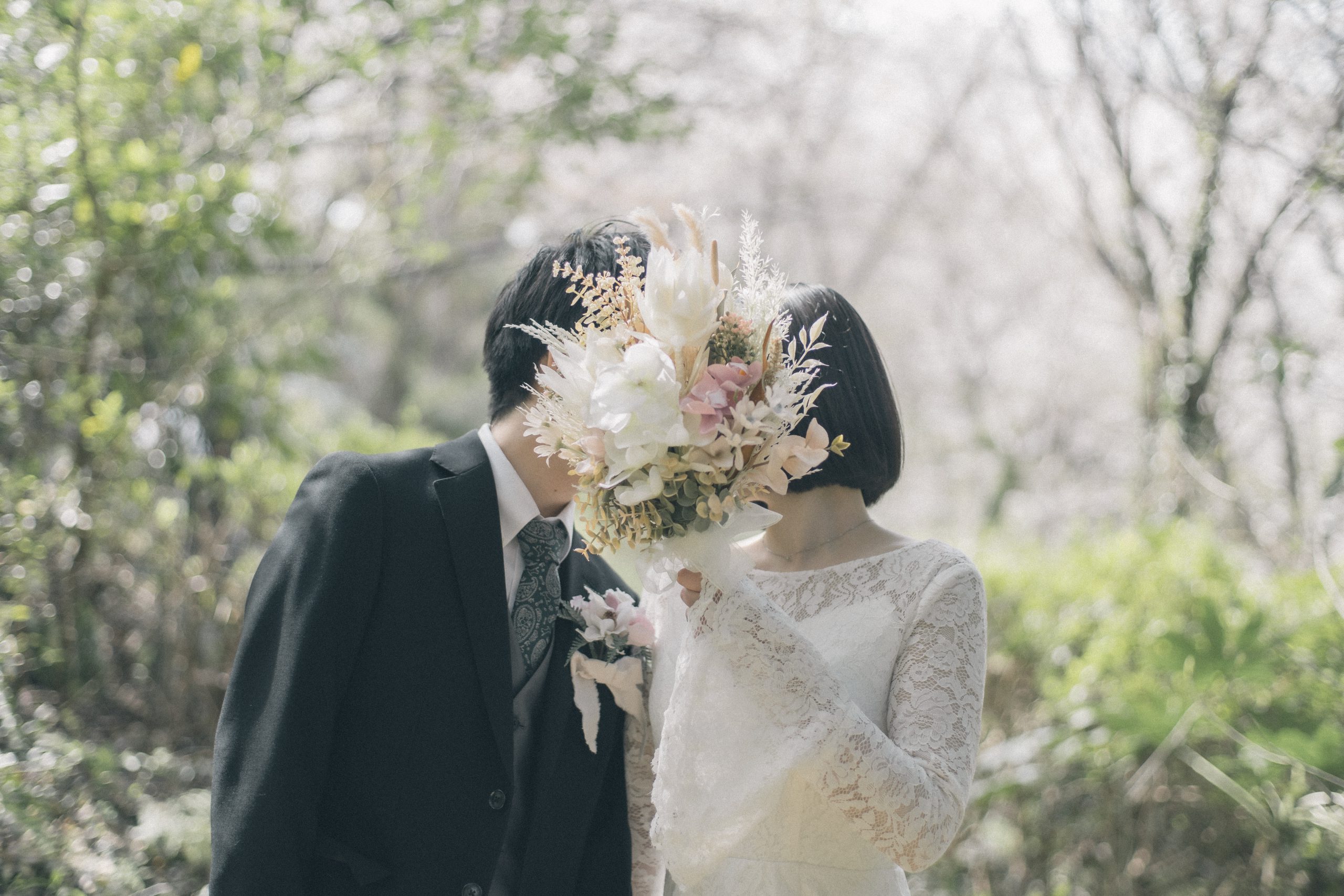 桜の下でブーケ越しに顔を寄せる新郎新婦