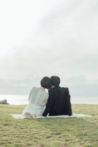 浜辺の芝生に並んで座り海を眺める新郎新婦の後ろ姿