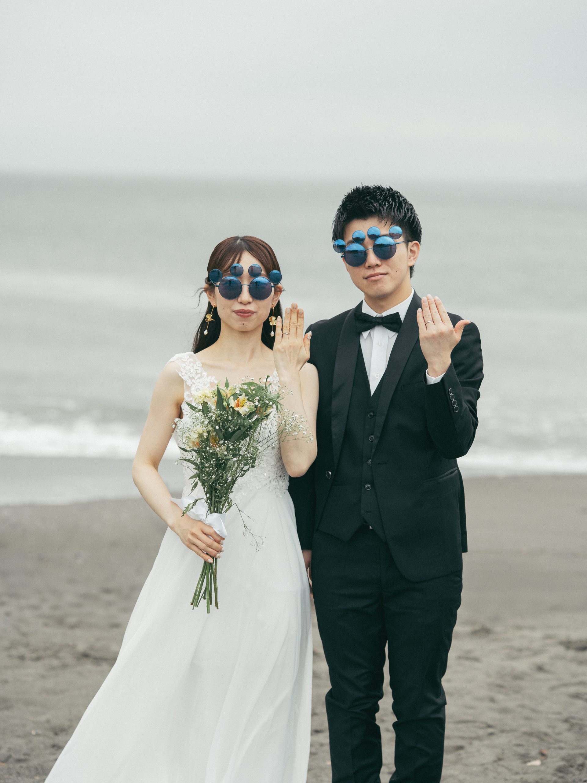 保田中央海岸で青い丸眼鏡をかけ指輪を見せる新郎新婦